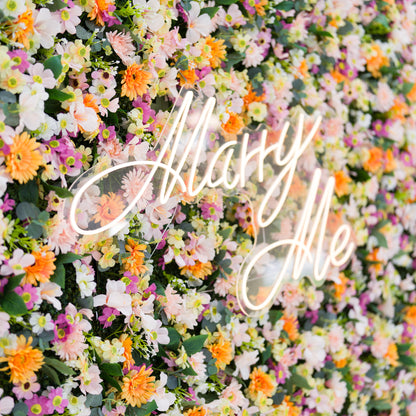 This close-up features a lush floral backdrop: dense clusters of warm orange daisies, soft pale pink blooms, crisp white flowers, and subtle purple accents, all anchored by a glowing "Marry Me" neon sign (cursive, light-lit). Lush green leaves add texture, while the bright pastel-and-orange palette evokes a romantic, cheerful vibe—ideal for proposal setups, wedding decor, or intimate event styling.