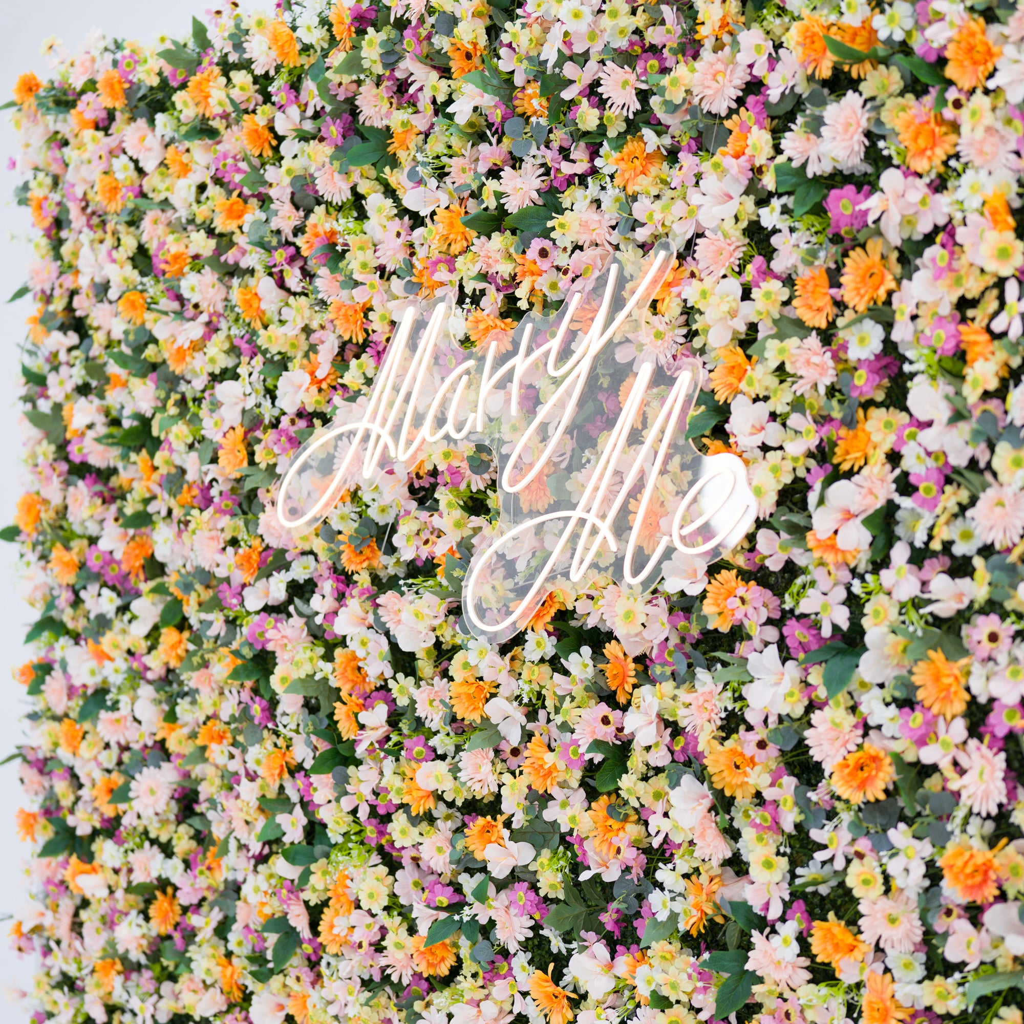 This vibrant floral backdrop bursts with warm orange daisies, soft pale pink blooms, crisp white flowers, and subtle purple accents, all densely clustered. A glowing "Marry Me" neon sign (cursive, light-lit) anchors the center, blending seamlessly with the lush petals. Lush green foliage adds texture, while the bright pastel-and-orange palette evokes a romantic, cheerful vibe—ideal for proposal setups, wedding decor, or intimate event styling.