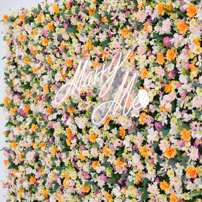 This vibrant floral backdrop bursts with warm orange daisies, soft pale pink blooms, crisp white flowers, and subtle purple accents, all densely clustered. A glowing "Marry Me" neon sign (cursive, light-lit) anchors the center, blending seamlessly with the lush petals. Lush green foliage adds texture, while the bright pastel-and-orange palette evokes a romantic, cheerful vibe—ideal for proposal setups, wedding decor, or intimate event styling.