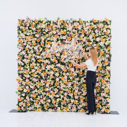 This square floral backdrop (against a plain white background) bursts with warm orange daisies, soft pale pink blooms, crisp white flowers, and subtle purple accents. A glowing "Marry Me" neon sign anchors the center, while a woman (white top, black wide-leg pants, heels) adjusts it, adding a dynamic touch. The bright, dense blooms evoke a romantic, cheerful vibe—ideal for proposal setups, wedding decor, or intimate event styling.