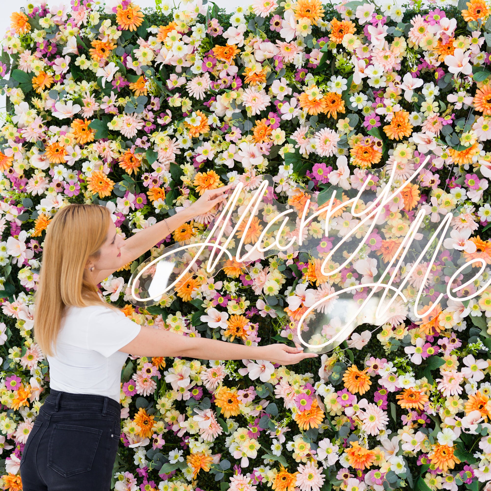 This close-up captures a woman (white top, black jeans) adjusting a glowing "Marry Me" neon sign, set against a dense floral backdrop of warm orange daisies, soft pale pink blooms, crisp white flowers, and subtle purple accents. Lush green foliage frames the vibrant petals, while the bright, romantic palette evokes a cheerful, intimate vibe—ideal for proposal setups, wedding decor, or heartfelt event styling.