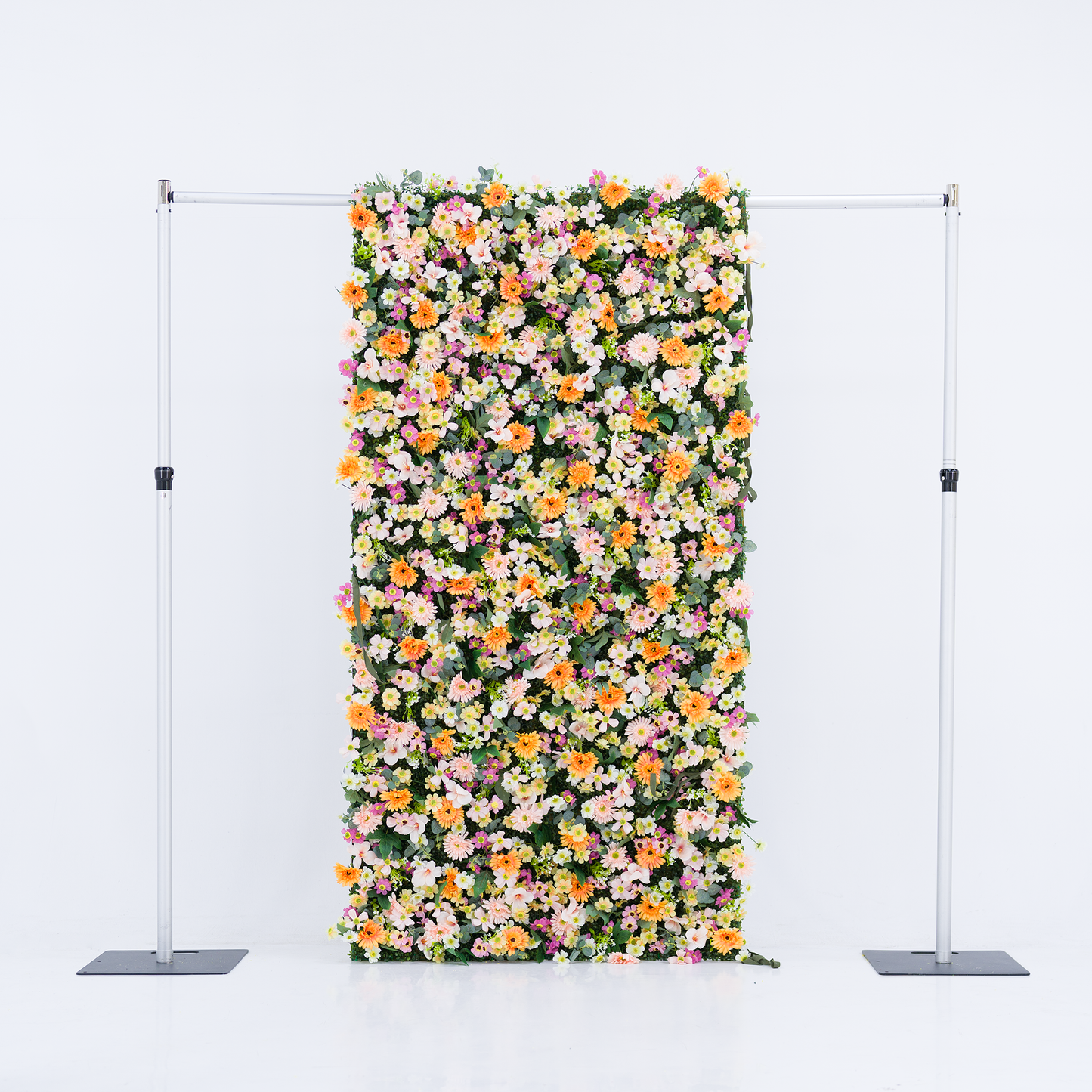This vertical floral backdrop (mounted on a sleek silver frame, set against a plain white background) bursts with dense clusters of warm orange daisies, soft pale pink blooms, crisp white flowers, and subtle purple accents. Lush green foliage weaves through the vibrant petals, creating a full, cheerful display. The bright pastel-and-orange palette evokes a fresh, lively vibe—ideal for event backdrops, photo booths, or festive decor setups.