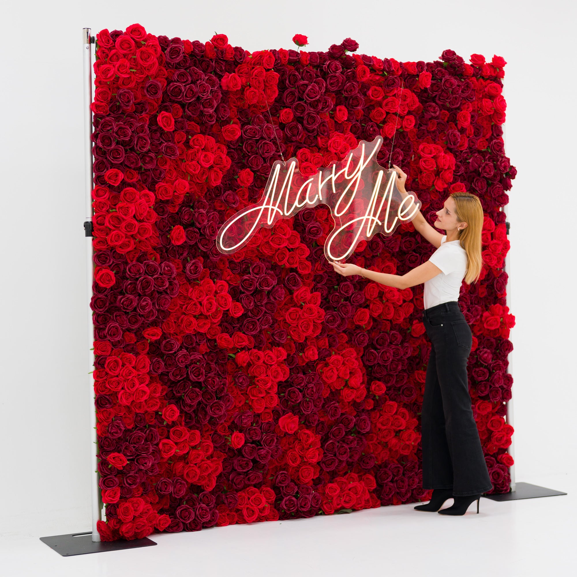 This vivid backdrop (against a plain white surface) is a dense wall of layered red and burgundy roses, radiating romantic lushness. A woman (white top, black wide-leg pants, heels) adjusts a glowing "Marry Me" neon sign—its cursive light contrasting beautifully with the deep blooms. The bold, rich palette exudes passion and elegance, perfect for proposal setups, romantic events, or wedding decor.