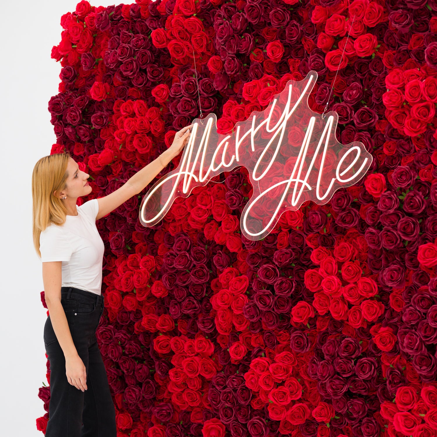 This close-up captures a woman (white top, black jeans) adjusting a glowing "Marry Me" neon sign, set against a dense wall of rich red and burgundy roses. The sign’s cursive light contrasts vividly with the lush, layered petals, exuding romantic warmth. The bold, elegant palette makes this scene perfect for proposal setups, wedding decor, or intimate romantic events.