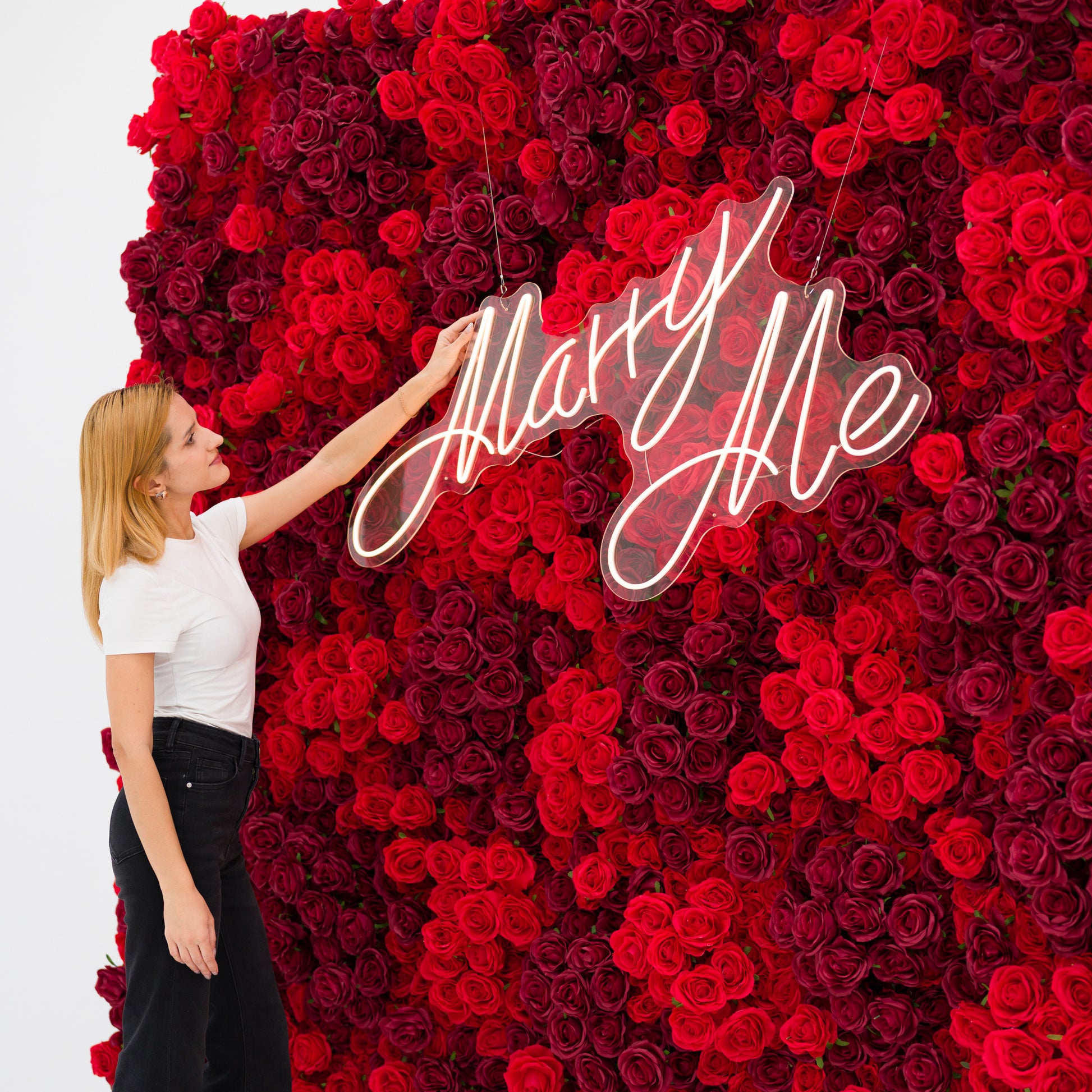 This close-up captures a woman (white top, black jeans) adjusting a glowing "Marry Me" neon sign, set against a dense wall of rich red and burgundy roses. The sign’s cursive light contrasts vividly with the lush, layered petals, exuding romantic warmth. The bold, elegant palette makes this scene perfect for proposal setups, wedding decor, or intimate romantic events.