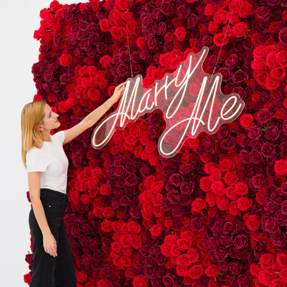 This close-up captures a woman (white top, black jeans) adjusting a glowing "Marry Me" neon sign, set against a dense wall of rich red and burgundy roses. The sign’s cursive light contrasts vividly with the lush, layered petals, exuding romantic warmth. The bold, elegant palette makes this scene perfect for proposal setups, wedding decor, or intimate romantic events.