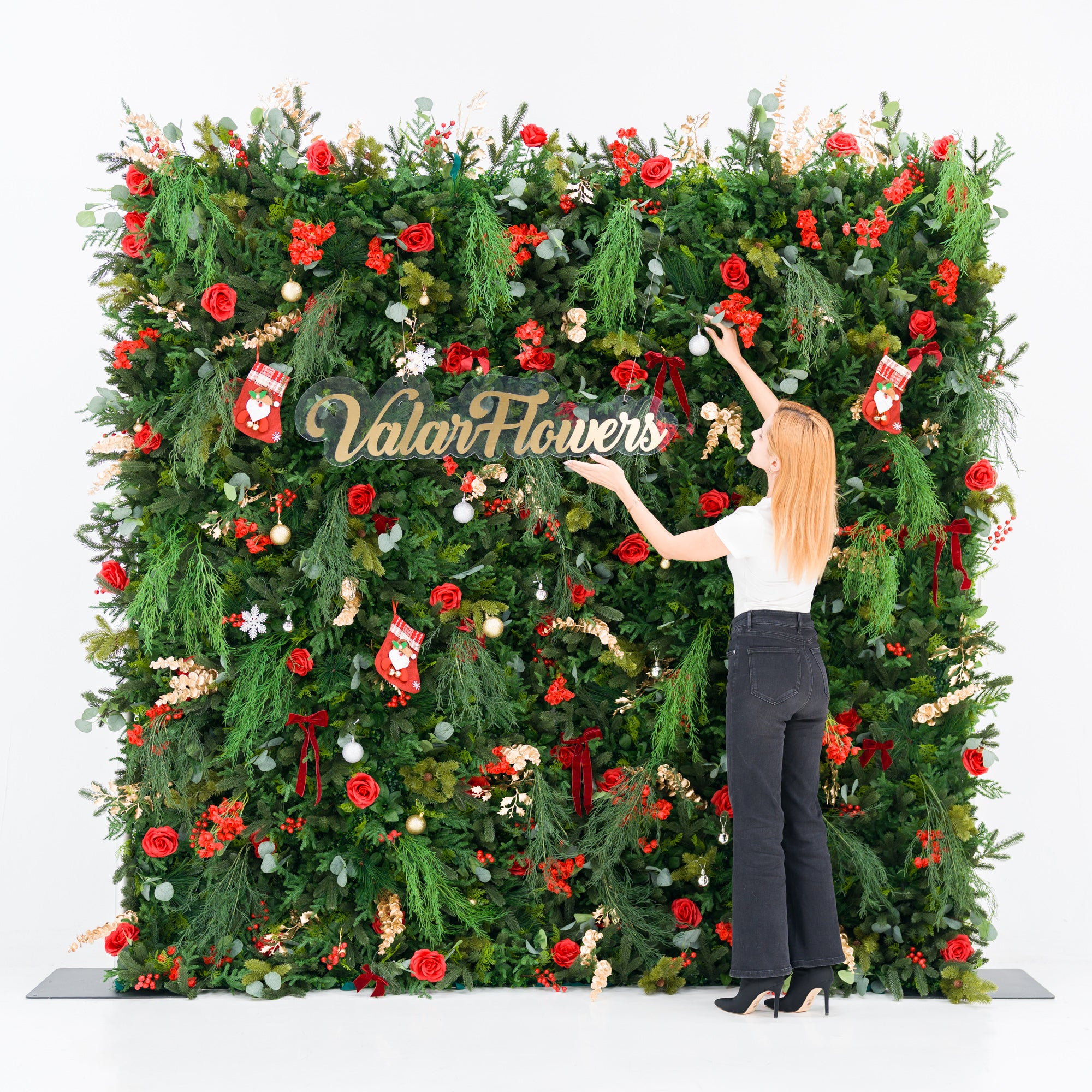 This festive square backdrop (against a plain white background) is lush with green foliage, red roses, clustered blooms, and red berries, accented by mini Santa stockings, gold/silver ornaments, and a polished “ValarFlowers” sign. A woman (white top, black wide-leg jeans, heels) adjusts a white ornament, adding a dynamic styling touch. The rich red-and-green palette blends cozy holiday charm with elegant texture—ideal for seasonal events or festive decor setups.