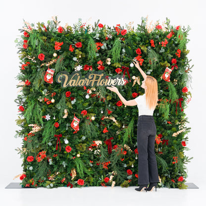 This festive square backdrop (against a plain white background) is lush with green foliage, red roses, clustered blooms, and red berries, accented by mini Santa stockings, gold/silver ornaments, and a polished “ValarFlowers” sign. A woman (white top, black wide-leg jeans, heels) adjusts a white ornament, adding a dynamic styling touch. The rich red-and-green palette blends cozy holiday charm with elegant texture—ideal for seasonal events or festive decor setups.