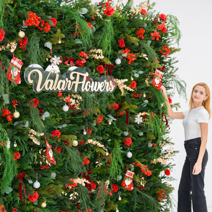 This lush festive greenery wall (against a plain white background) is dense with pine/feathery foliage, red roses, and red berry clusters. Accents include mini Santa stockings, gold/silver ornaments, and gold sprays, plus a polished “ValarFlowers” sign. A woman (white top, black jeans) adjusts a stocking, adding a dynamic styling touch. The rich red-and-green palette blends cozy holiday charm with elegant texture—ideal for seasonal events or festive decor setups.
