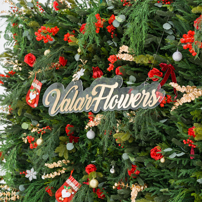 This close-up captures a lush festive arrangement: dense green foliage (pine, feathery fronds) mixed with bold red roses, clustered blooms, and red berries. Accents include mini Santa stockings, gold/silver ornaments, snowflakes, and a polished “ValarFlowers” sign. Varied textures—soft petals, lush greenery, shiny decor—blend with the rich red-and-green palette, evoking cozy holiday charm, perfect for seasonal decor or festive event styling.