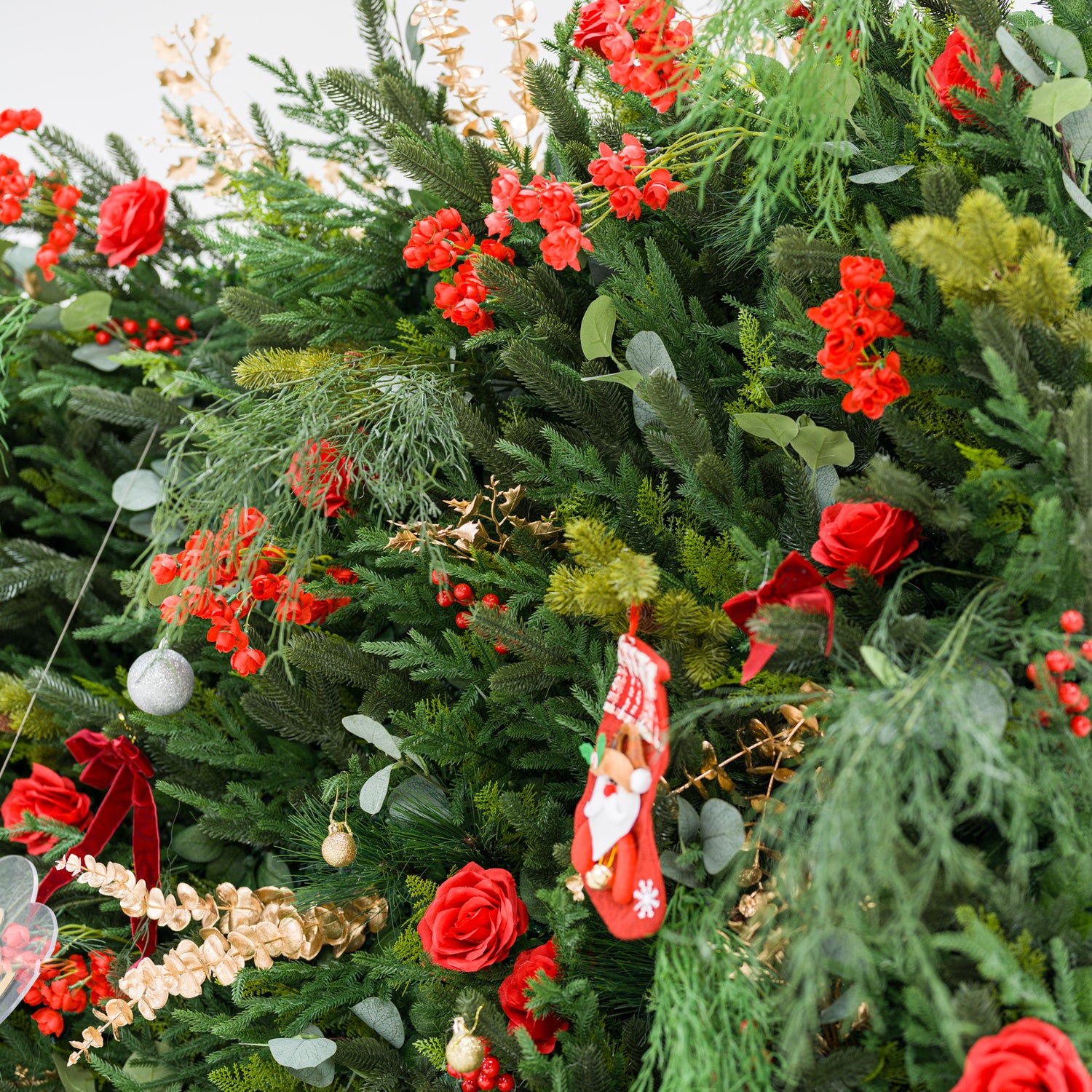 This close-up shows a lush festive arrangement: dense green foliage (pine, feathery fronds) mixed with bold red roses, clustered blooms, and red berries. Accents include a mini Santa stocking, gold/silver ornaments, a red velvet bow, and delicate gold sprays. Varied textures—soft petals, lush greenery, shiny decor—blend with the rich red-and-green palette, evoking cozy holiday charm, perfect for seasonal decor or festive event styling.