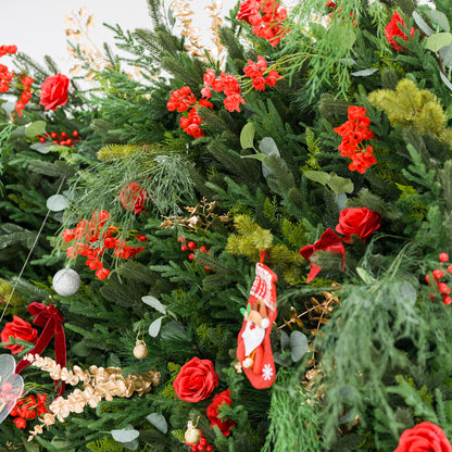 This close-up shows a lush festive arrangement: dense green foliage (pine, feathery fronds) mixed with bold red roses, clustered blooms, and red berries. Accents include a mini Santa stocking, gold/silver ornaments, a red velvet bow, and delicate gold sprays. Varied textures—soft petals, lush greenery, shiny decor—blend with the rich red-and-green palette, evoking cozy holiday charm, perfect for seasonal decor or festive event styling.
