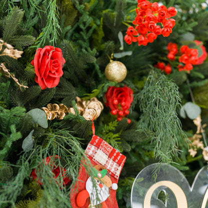This intimate close-up showcases a festive arrangement: dense green foliage (pine, feathery fronds) paired with bold red roses, clustered blooms, and red berries. Accents include a mini Santa stocking (plaid pattern, whimsical design), a shiny gold ornament, and delicate gold sprays. Varied textures—soft petals, lush greenery, textured decor—blend with the rich red-and-green palette, evoking cozy holiday charm, perfect for seasonal decor or festive styling details.