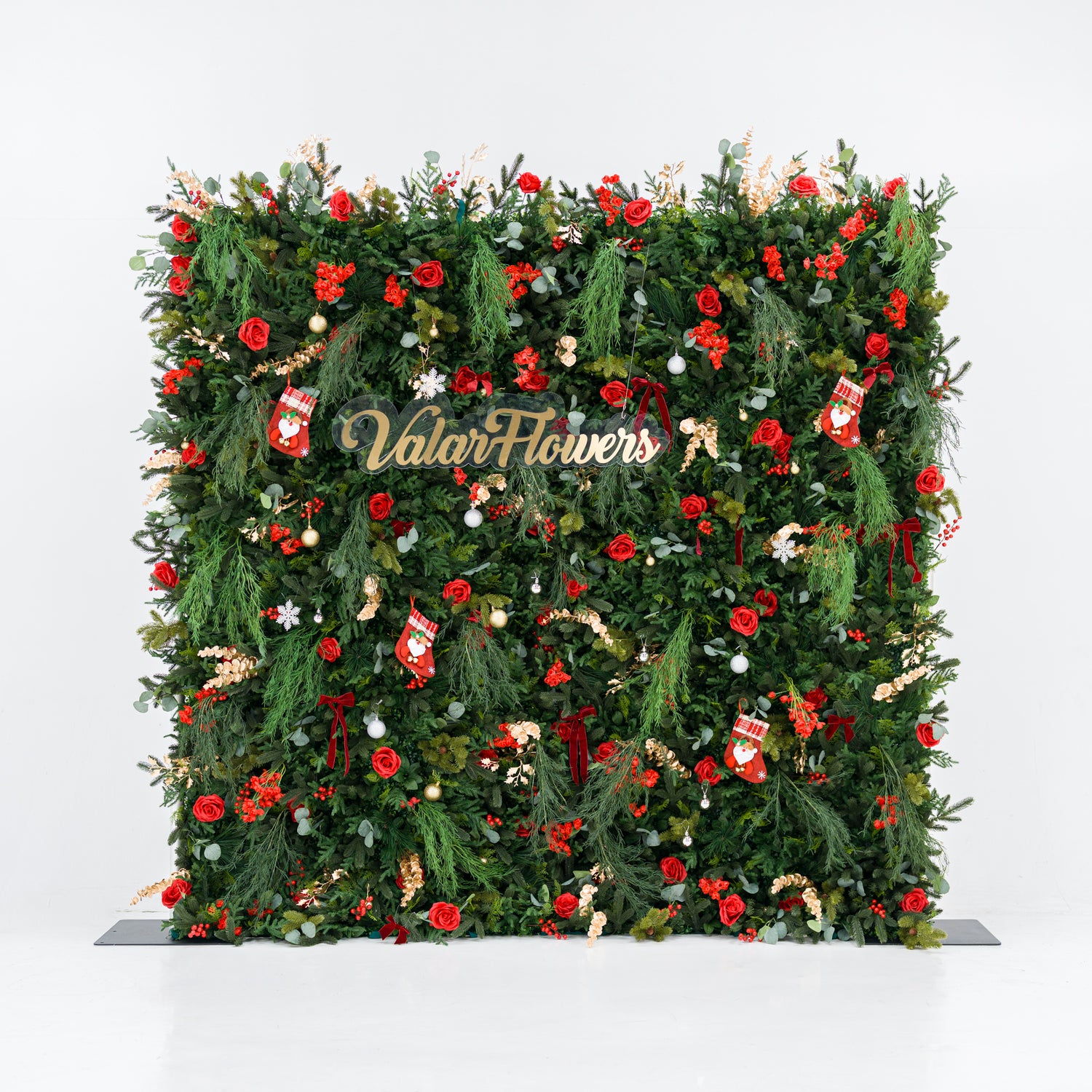 This square festive backdrop (against a plain white background) is densely covered with lush green foliage, bold red roses, clustered red blooms, and berry clusters. Accents include mini Santa stockings, gold/silver ornaments, snowflakes, and a polished "ValarFlowers" sign. The rich red-and-green palette, layered textures, and cohesive styling evoke cozy holiday charm—ideal for Christmas events, festive photo ops, or seasonal decor setups.