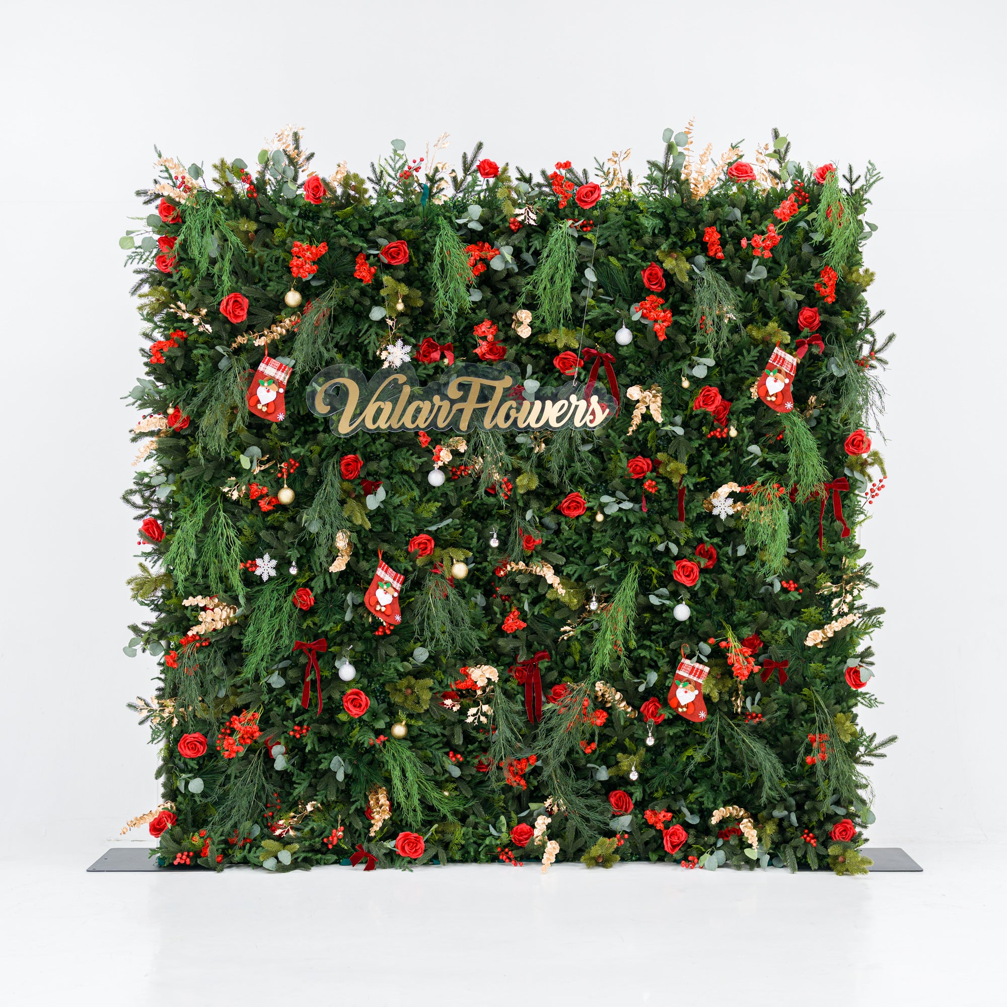 This square festive backdrop (against a plain white background) is densely covered with lush green foliage, bold red roses, clustered red blooms, and berry clusters. Accents include mini Santa stockings, gold/silver ornaments, snowflakes, and a polished "ValarFlowers" sign. The rich red-and-green palette, layered textures, and cohesive styling evoke cozy holiday charm—ideal for Christmas events, festive photo ops, or seasonal decor setups.