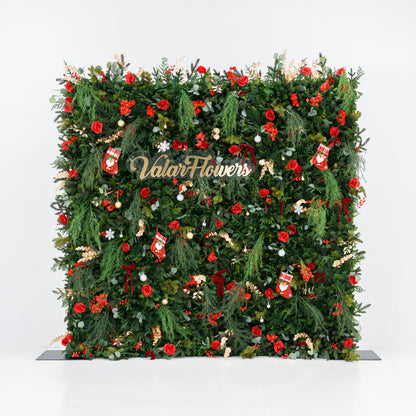 This square festive backdrop (against a plain white background) is densely covered with lush green foliage, bold red roses, clustered red blooms, and berry clusters. Accents include mini Santa stockings, gold/silver ornaments, snowflakes, and a polished "ValarFlowers" sign. The rich red-and-green palette, layered textures, and cohesive styling evoke cozy holiday charm—ideal for Christmas events, festive photo ops, or seasonal decor setups.