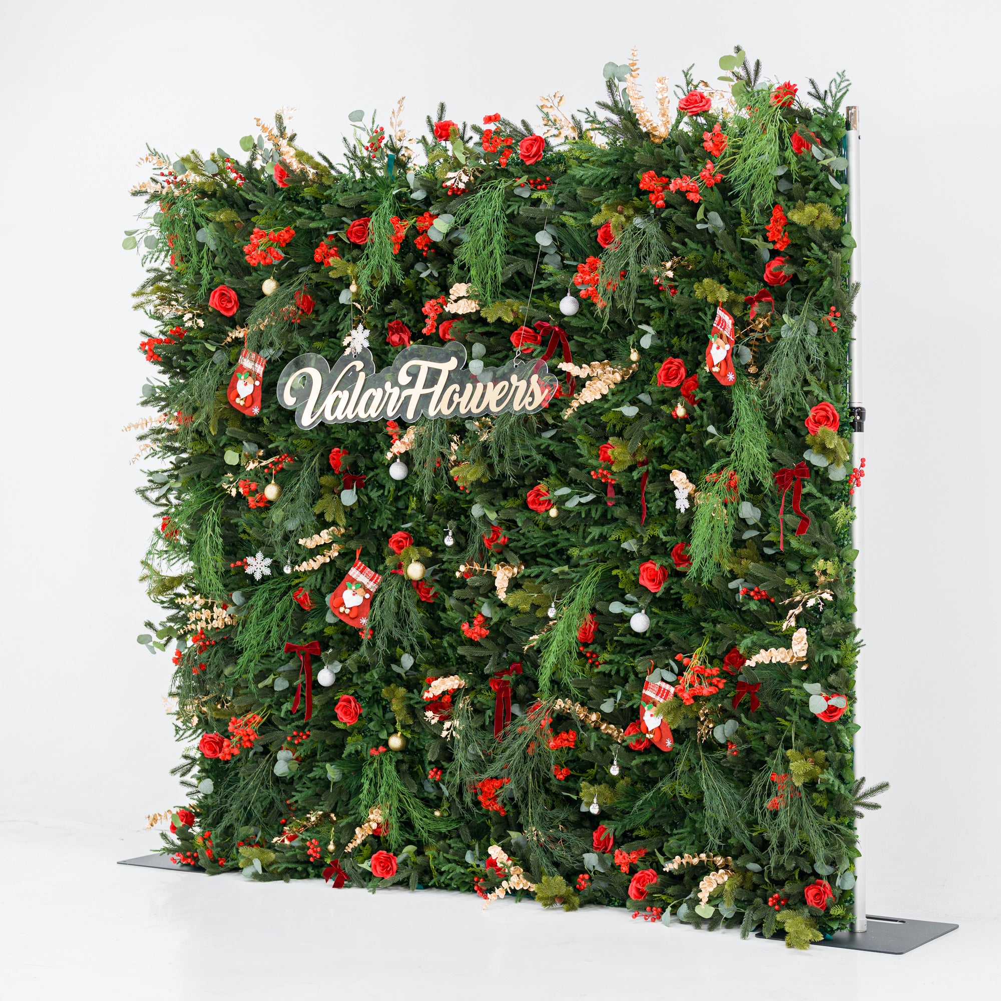 This tall, festive greenery backdrop (against a plain white background) is dense with lush foliage, bold red roses, clustered blooms, and berry clusters. Accents include mini Santa stockings, gold/silver ornaments, snowflakes, and a polished "ValarFlowers" sign. A metallic frame edge peeks through, grounding the vibrant design. The rich red-and-green palette blends cozy holiday charm with elegant texture—ideal for seasonal events or festive decor setups.