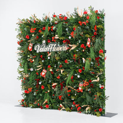 This tall, festive greenery backdrop (against a plain white background) is dense with lush foliage, bold red roses, clustered blooms, and berry clusters. Accents include mini Santa stockings, gold/silver ornaments, snowflakes, and a polished "ValarFlowers" sign. A metallic frame edge peeks through, grounding the vibrant design. The rich red-and-green palette blends cozy holiday charm with elegant texture—ideal for seasonal events or festive decor setups.