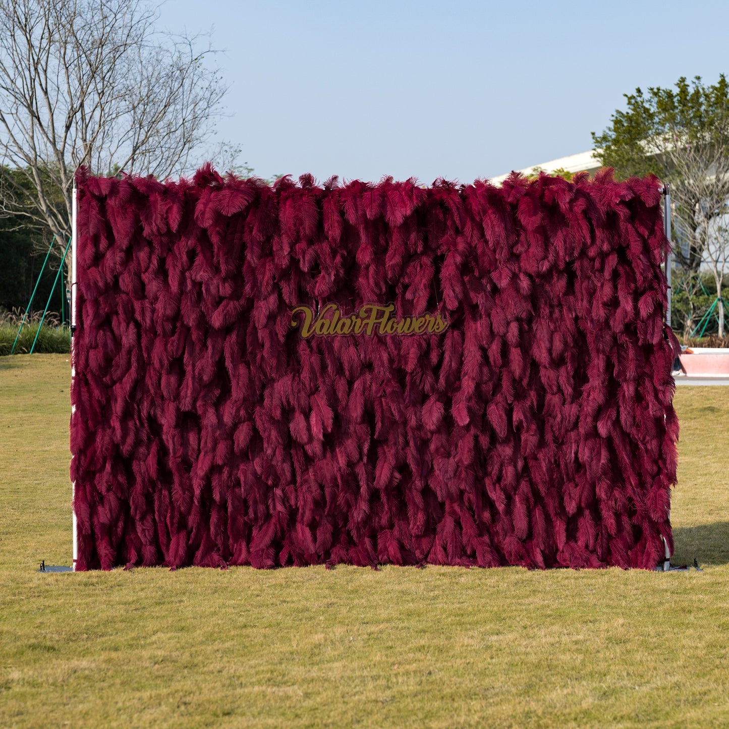 This striking outdoor backdrop features a dense, velvety wall of deep burgundy artificial foliage, with a soft, feathery texture that creates rich, vertical depth. Centered on the surface, a gold cursive logo reads “ValarFlowers,” adding a touch of refined elegance. It stands on a sunlit green lawn, framed by trees under a clear sky, making it a luxurious, eye-catching focal point for weddings, brand activations, or upscale outdoor events.