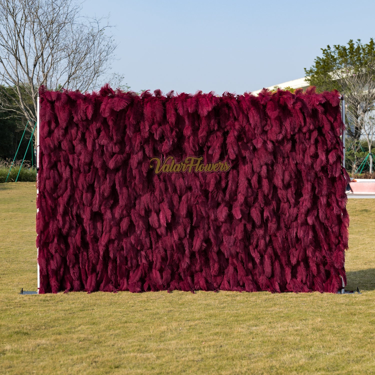 This striking outdoor backdrop features a dense, velvety wall of deep burgundy artificial foliage, with a soft, feathery texture that creates rich, vertical depth. Centered on the surface, a gold cursive logo reads “ValarFlowers,” adding a touch of refined elegance. It stands on a sunlit green lawn, framed by trees under a clear sky, making it a luxurious, eye-catching focal point for weddings, brand activations, or upscale outdoor events.