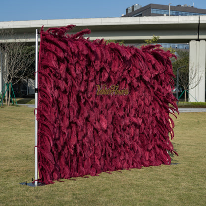This opulent 12x8ft (3.6x2.4m) outdoor backdrop features a dense wall of deep burgundy ostrich feathers, creating a plush, velvety texture with rich vertical depth. Centered on its surface, a glossy gold cursive logo reads “ValarFlowers,” adding a refined, elegant touch. It stands on a sunlit green lawn, framed by modern architecture and trees, making it a striking focal point for weddings, brand activations, or upscale outdoor events.