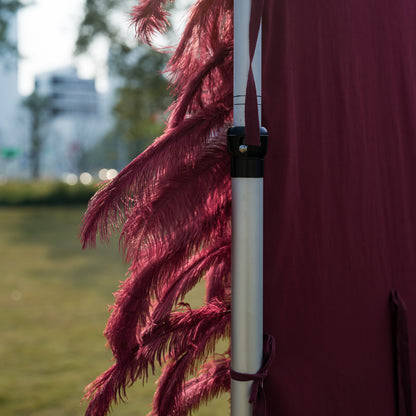 This elegant outdoor event detail features a cascading fringe of deep burgundy ostrich feathers, their soft, wispy plumes spilling over the edge of a sleek, maroon fabric backdrop mounted on a silver metal frame. Set against a sunlit lawn, distant trees, and modern buildings, the plush, velvety texture of the feathers contrasts beautifully with the crisp fabric, creating a sophisticated, romantic touch for weddings, proposals, or upscale gatherings.
