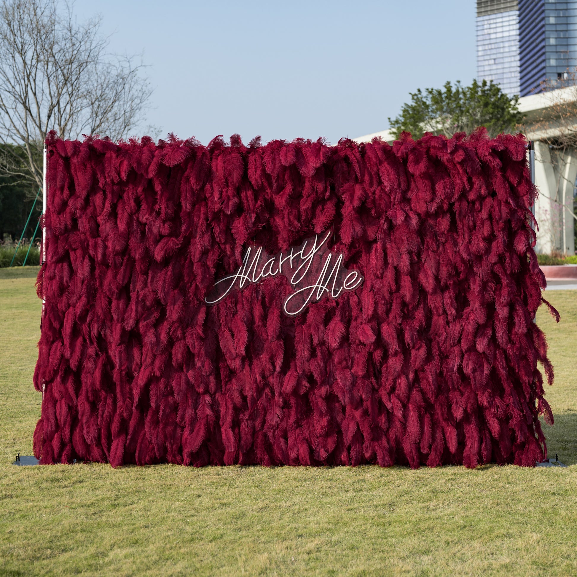 This luxurious outdoor proposal backdrop features a dense wall of deep burgundy ostrich feathers, creating a plush, velvety texture with rich vertical depth. At its center, a sleek, clear acrylic sign reads “Marry Me” in elegant white cursive, adding a romantic, modern touch. Set against a sunlit green lawn and framed by contemporary architecture, it is an opulent focal point for unforgettable proposals.
