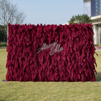 This luxurious outdoor proposal backdrop features a dense wall of deep burgundy ostrich feathers, creating a plush, velvety texture with rich vertical depth. At its center, a sleek, clear acrylic sign reads “Marry Me” in elegant white cursive, adding a romantic, modern touch. Set against a sunlit green lawn and framed by contemporary architecture, it is an opulent focal point for unforgettable proposals.
