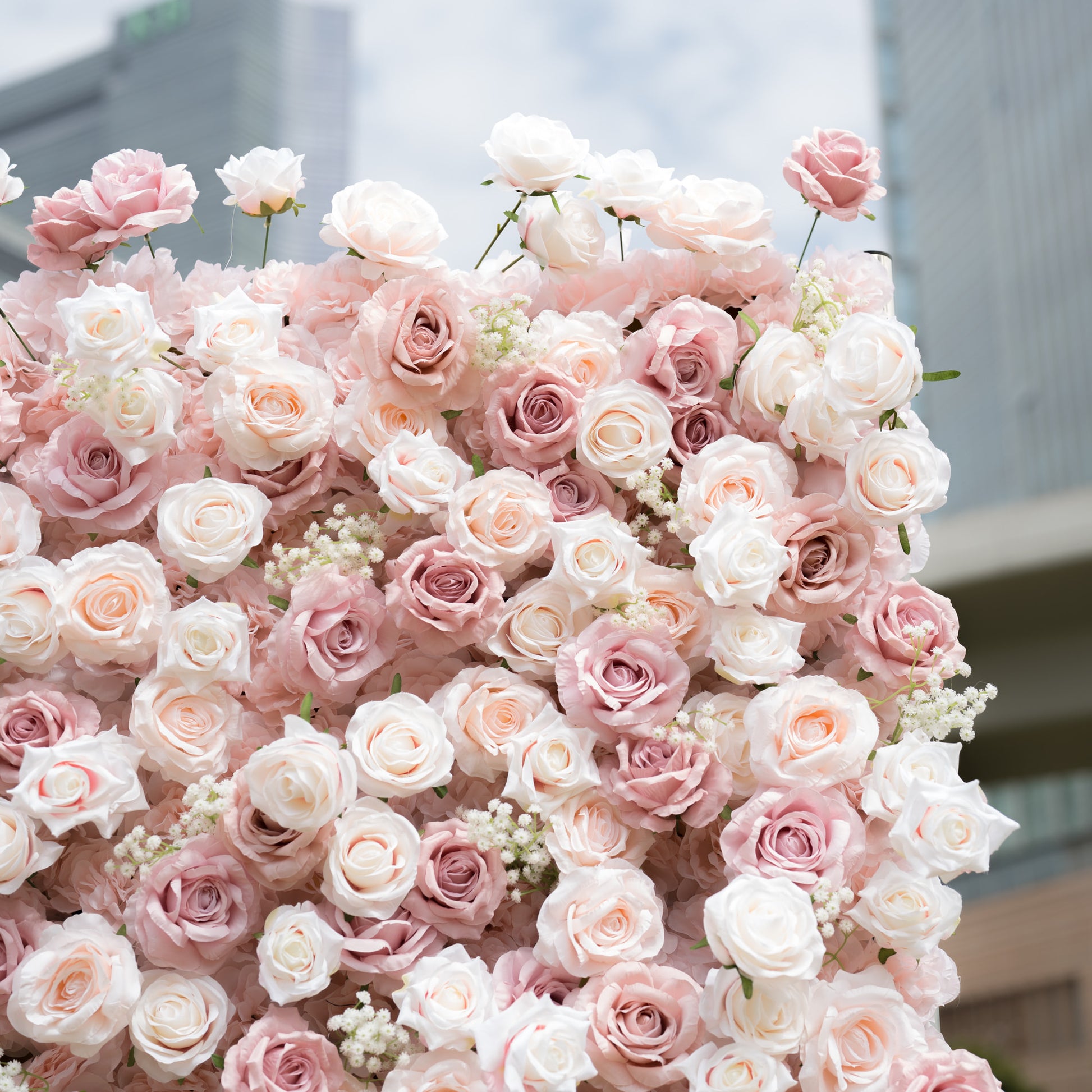 This Valar Flowers floral wall is a lush, romantic display of creamy white, soft blush, and dusty pink roses, interspersed with delicate baby's breath. Set against a modern cityscape, its dense, layered blooms create a dreamy, elegant texture, ideal for luxury wedding backdrops, photo shoots, and high-end event decor.