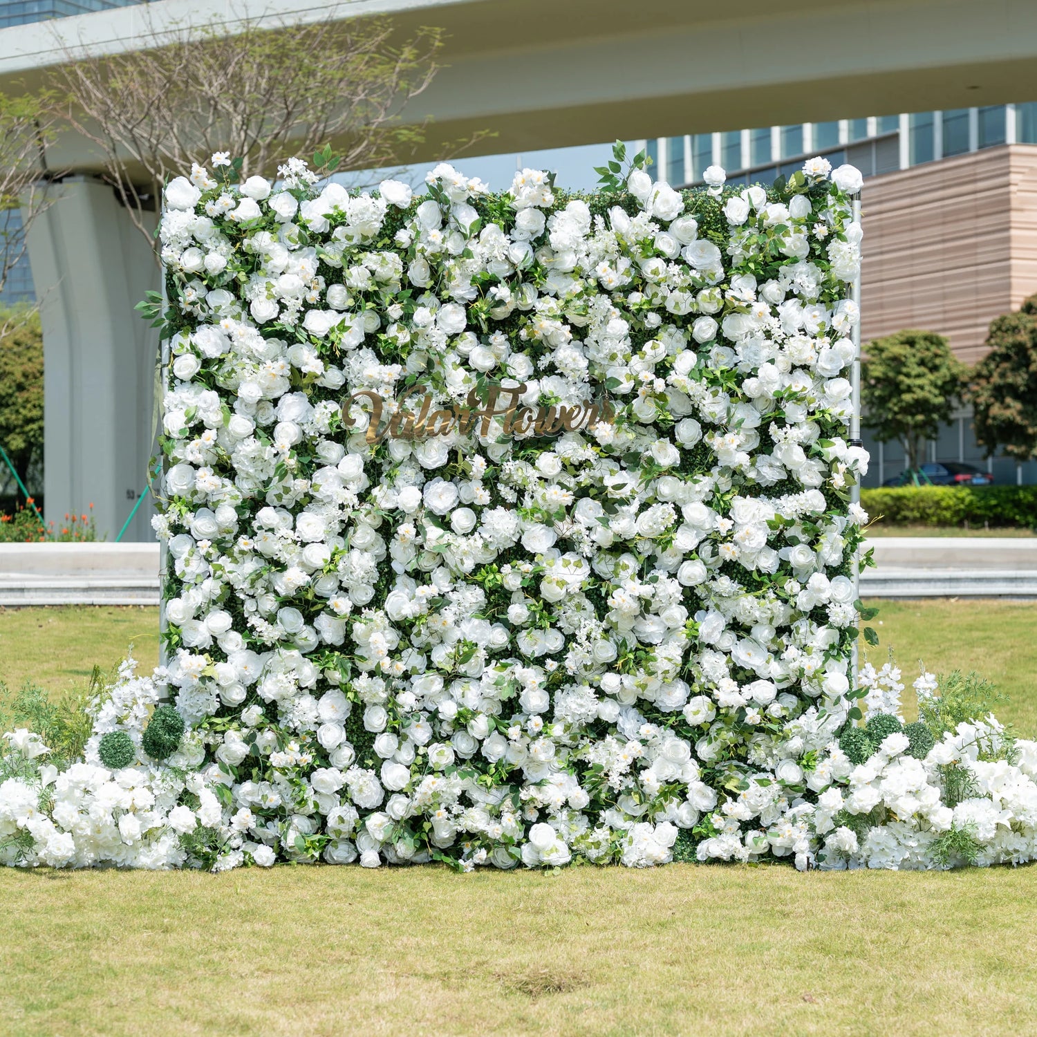 This is a premium all-white artificial flower wall backdrop by Valar Flowers, featuring a lush, dense design of soft roses, peonies, delicate baby’s breath, and subtle green foliage. Centered with the metallic "ValarFlowers" logo, this reusable, maintenance-free display creates an elegant, timeless focal point. Ideal for outdoor weddings, photo booths, luxury events, and brand activations, it offers a clean, romantic aesthetic perfect for professional styling.