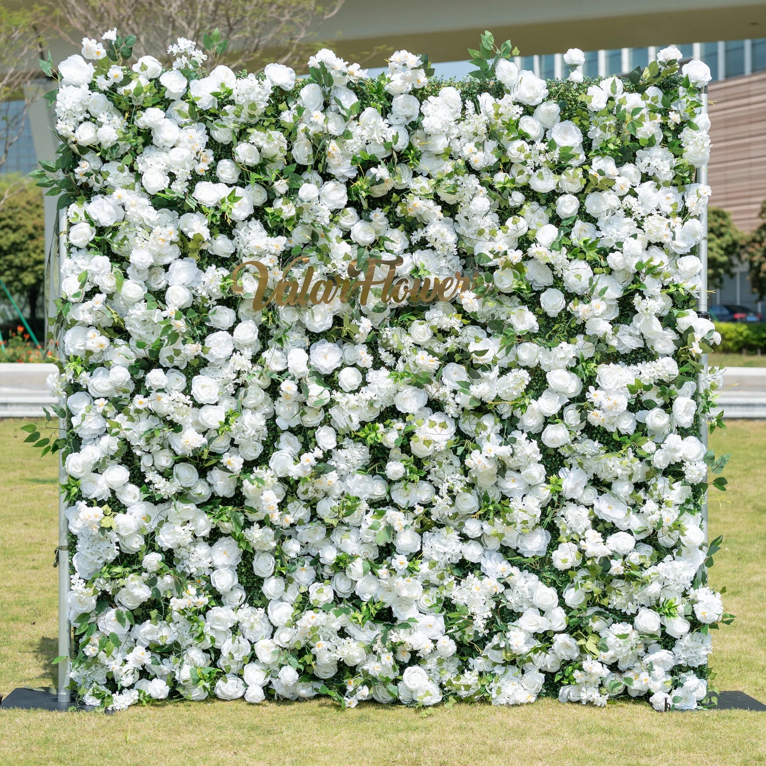 This is a premium all-white artificial flower wall backdrop by Valar Flowers, featuring a lush, dense design of soft roses, peonies, delicate baby’s breath, and subtle green foliage. Centered with the metallic "ValarFlowers" logo, this reusable, maintenance-free display creates an elegant, timeless focal point. Ideal for outdoor weddings, photo booths, luxury events, and brand activations, it offers a clean, romantic aesthetic perfect for professional styling.