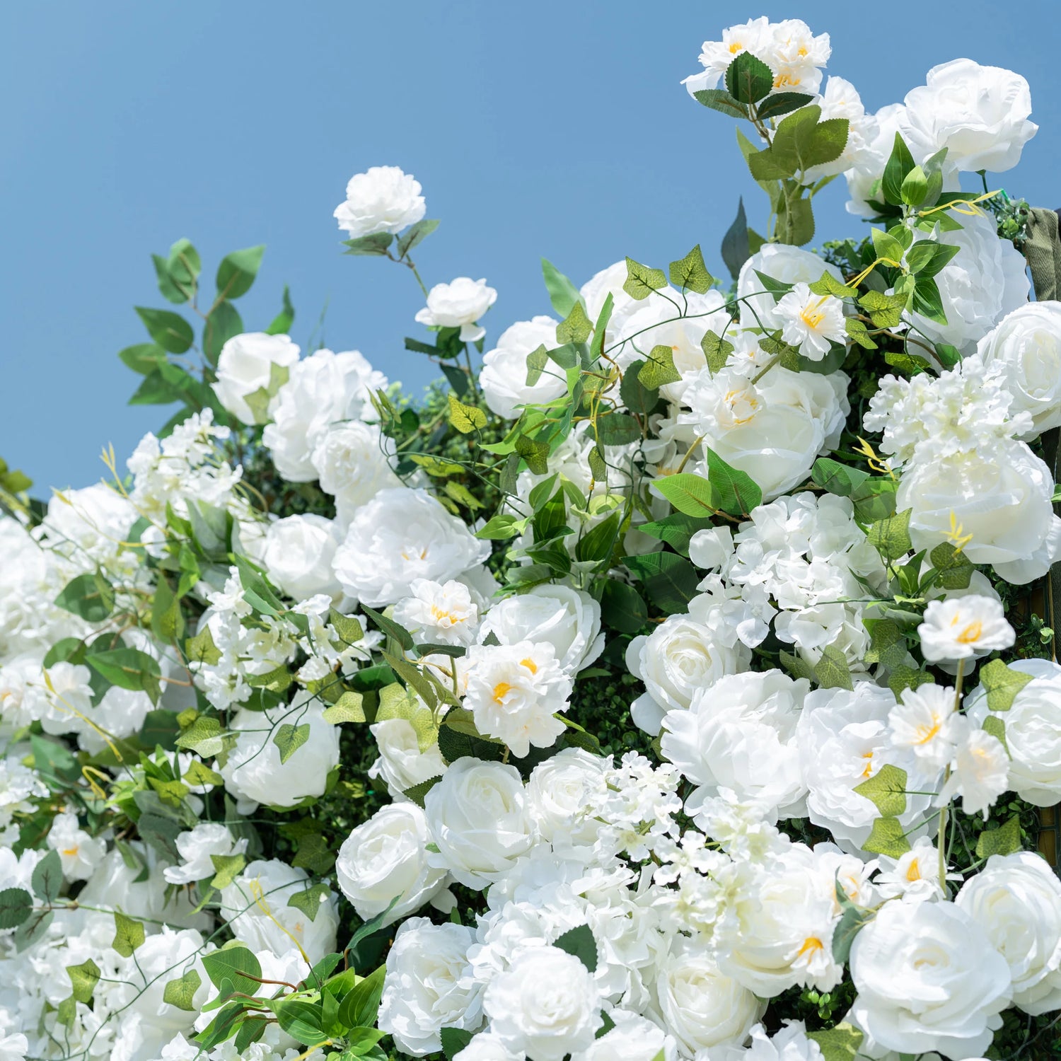 This close-up showcases the premium craftsmanship of Valar Flowers’ all-white artificial floral wall, featuring lush, hyper-realistic roses, peonies, delicate hydrangeas, and vibrant green foliage. Soft-textured petals mimic fresh blooms, while dense, layered leaves create a lifelike, maintenance-free display. Ideal for weddings, photo booths, and luxury events, this durable, reusable decor offers a clean, romantic aesthetic perfect for professional styling.