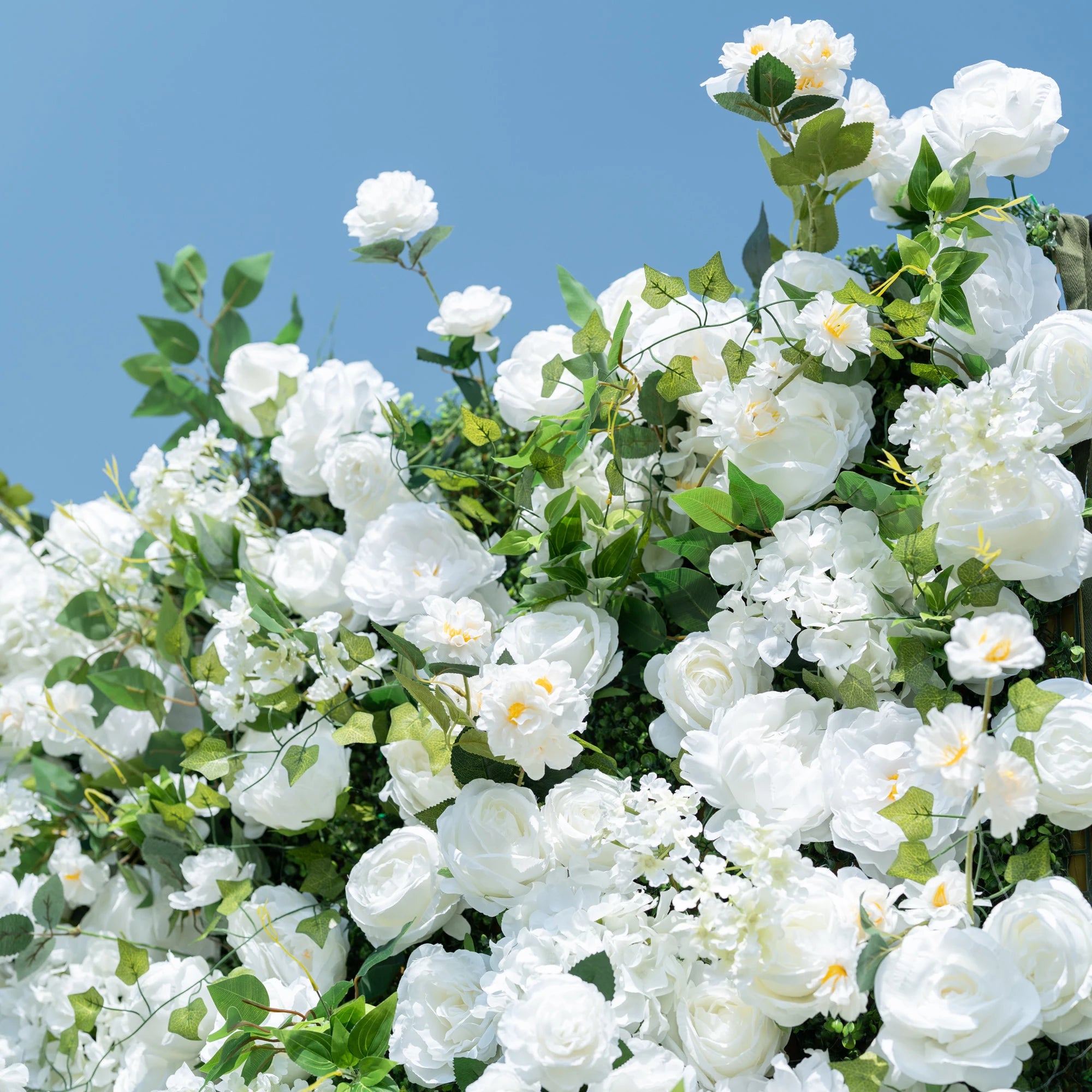 This close-up showcases the premium craftsmanship of Valar Flowers’ all-white artificial floral wall, featuring lush, hyper-realistic roses, peonies, delicate hydrangeas, and vibrant green foliage. Soft-textured petals mimic fresh blooms, while dense, layered leaves create a lifelike, maintenance-free display. Ideal for weddings, photo booths, and luxury events, this durable, reusable decor offers a clean, romantic aesthetic perfect for professional styling.