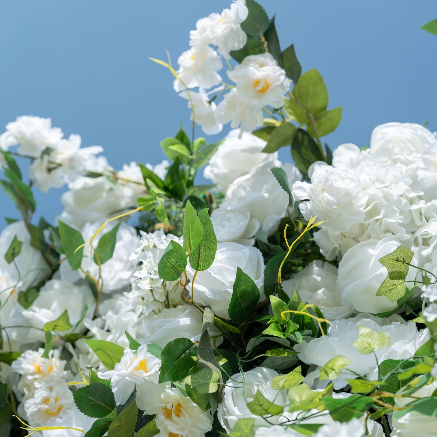This close-up highlights the premium craftsmanship of Valar Flowers’ artificial floral arrangement, featuring lush white roses, peonies, and delicate filler blooms paired with vibrant, realistic green foliage. The soft-textured petals mimic fresh flowers, while layered leaves and subtle yellow accents create a lifelike, maintenance-free display. Ideal for weddings, photo booths, and luxury events, this durable decor offers a clean, romantic aesthetic perfect for professional styling.