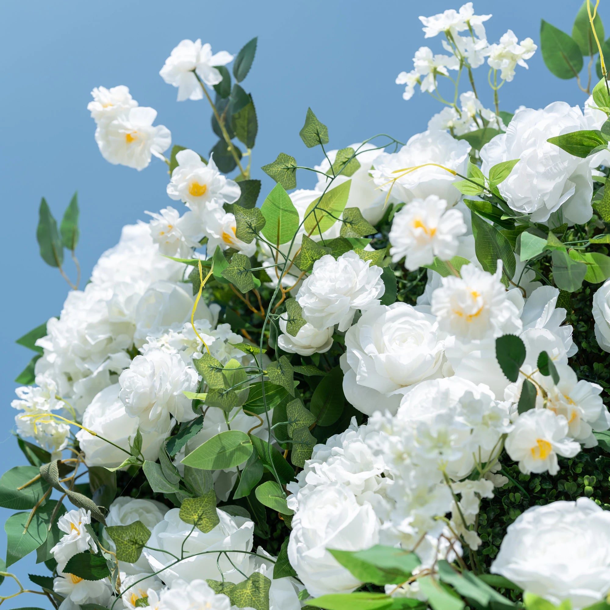 This close-up showcases Valar Flowers’ premium artificial white floral arrangement, featuring lush, hyper-realistic roses, peonies, and delicate filler blooms paired with vibrant green foliage. Soft-textured petals mimic fresh flowers, while layered leaves and subtle yellow accents create a lifelike, maintenance-free display against a clear blue sky. Ideal for weddings, photo booths, and luxury events, this durable decor offers a clean, romantic aesthetic perfect for professional styling.