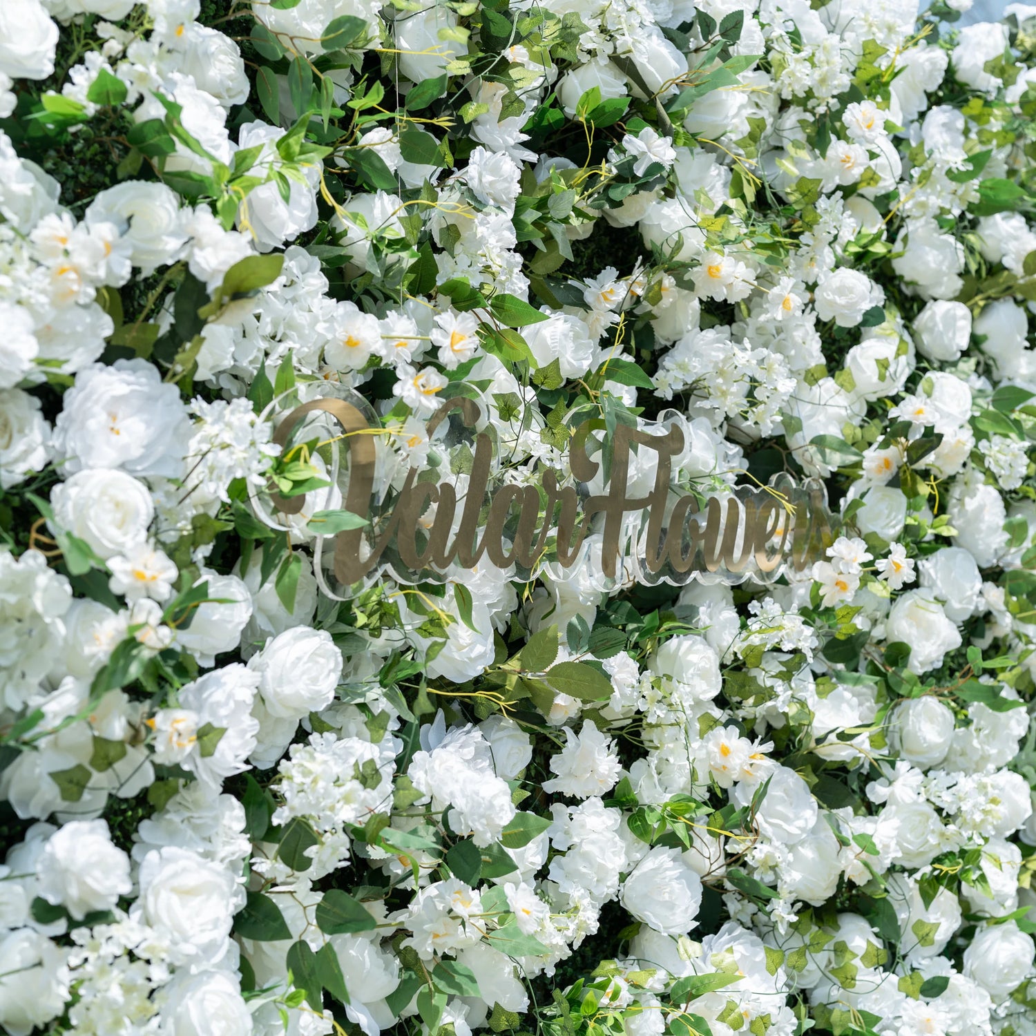 This close-up showcases Valar Flowers’ premium artificial white flower wall, featuring lush, hyper-realistic roses, peonies, and delicate filler blooms paired with vibrant green foliage. Centered with the metallic "ValarFlowers" logo, the dense, layered design highlights lifelike petals and soft textures, creating an elegant, maintenance-free display. Ideal for weddings, photo booths, and luxury events, this durable decor offers a clean, romantic aesthetic perfect for professional styling.