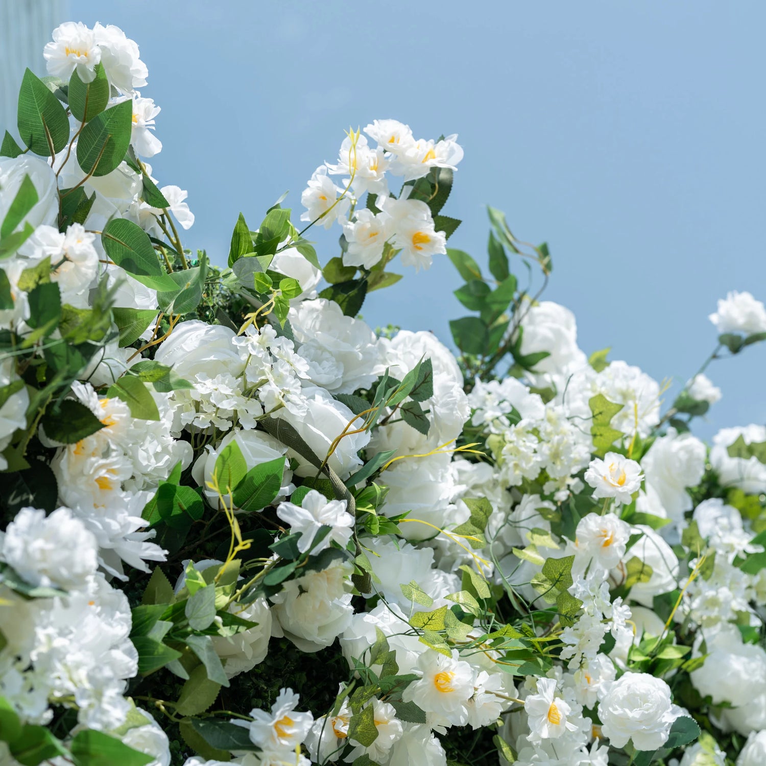This close-up captures the lush top edge of Valar Flowers’ premium artificial white floral wall, set against a soft light blue sky. Featuring hyper-realistic roses, peonies, and delicate filler blooms paired with vibrant green foliage, the layered design creates a romantic, airy aesthetic. Soft-textured petals and lifelike leaves mimic fresh flowers, offering a maintenance-free, durable display ideal for weddings, photo booths, and luxury events.