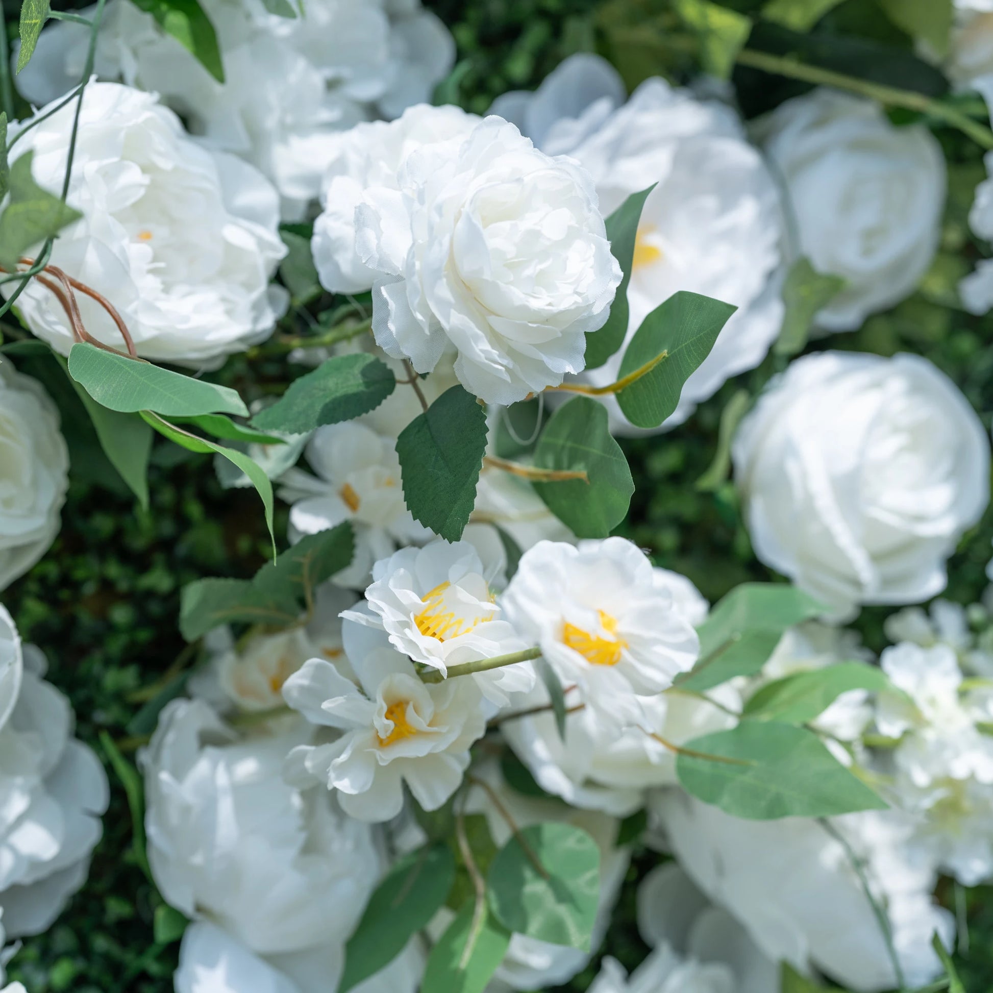 This close-up showcases the premium details of Valar Flowers’ artificial white floral wall, featuring lush, hyper-realistic roses, peonies, and delicate yellow-centered filler blooms paired with vibrant green foliage. Soft-textured petals mimic fresh flowers, while layered leaves and stems create a lifelike, maintenance-free display. Ideal for weddings, photo booths, and luxury events, this durable decor offers a clean, romantic aesthetic perfect for professional styling.