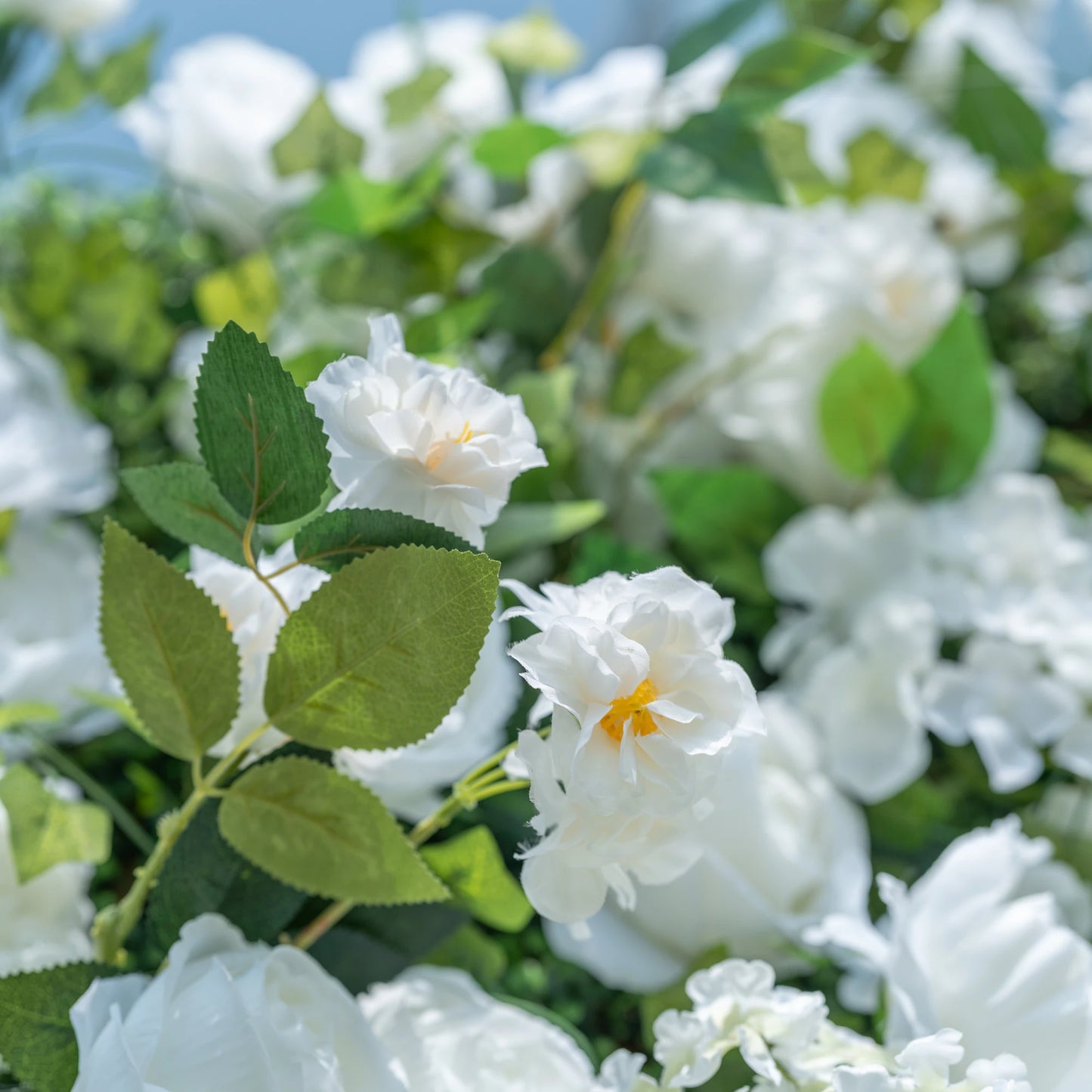 This close-up highlights Valar Flowers’ premium artificial white blooms, featuring delicate layered petals with soft yellow centers and hyper-realistic textured green foliage. The shallow depth of field emphasizes the intricate details of the flowers, creating a lifelike, romantic aesthetic. Maintenance-free and durable, these blooms are ideal for weddings, photo booths, and luxury events, offering a clean, timeless look perfect for professional styling.