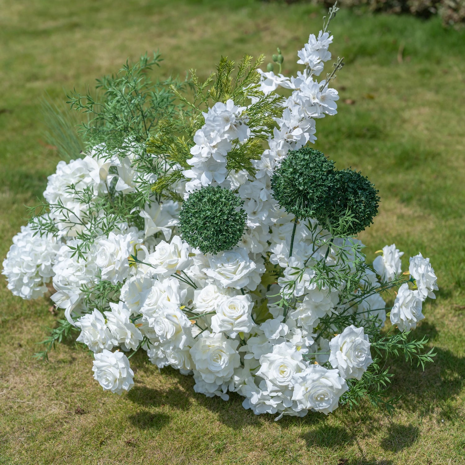 This premium artificial floral arrangement by Valar Flowers features a lush mix of white roses, hydrangeas, lilies, delphiniums, and textured green foliage, including spherical green accents. Designed for weddings, photo booths, and luxury events, this dense, realistic display offers a clean, timeless aesthetic. Maintenance-free and durable, it brings a romantic, elegant touch to any setting, perfect for professional styling and event decor.