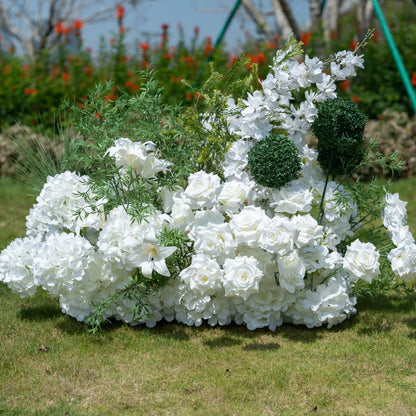 This full view showcases Valar Flowers’ premium artificial floral arrangement, featuring a lush mix of white roses, hydrangeas, lilies, delphiniums, and textured green foliage including spherical accents. Set against an outdoor garden backdrop, the dense, realistic display mimics fresh blooms, offering a clean, timeless aesthetic. Maintenance-free and durable, this elegant piece is ideal for weddings, photo booths, and luxury events, perfect for professional styling.