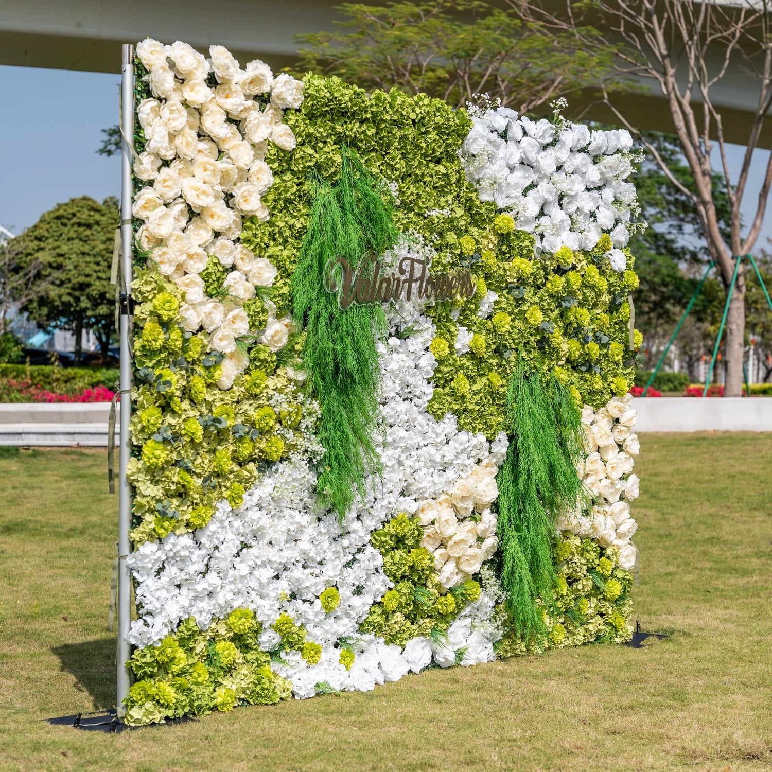This premium artificial flower wall backdrop by Valar Flowers features a lush, nature-inspired design with vibrant green hydrangeas, crisp white blooms, soft ivory roses, and cascading green foliage, accented by the custom "ValarFlowers" logo. Reusable and maintenance-free, it creates a fresh, elegant focal point ideal for outdoor weddings, garden parties, photo booths, and luxury events, offering long-lasting beauty for professional decor.