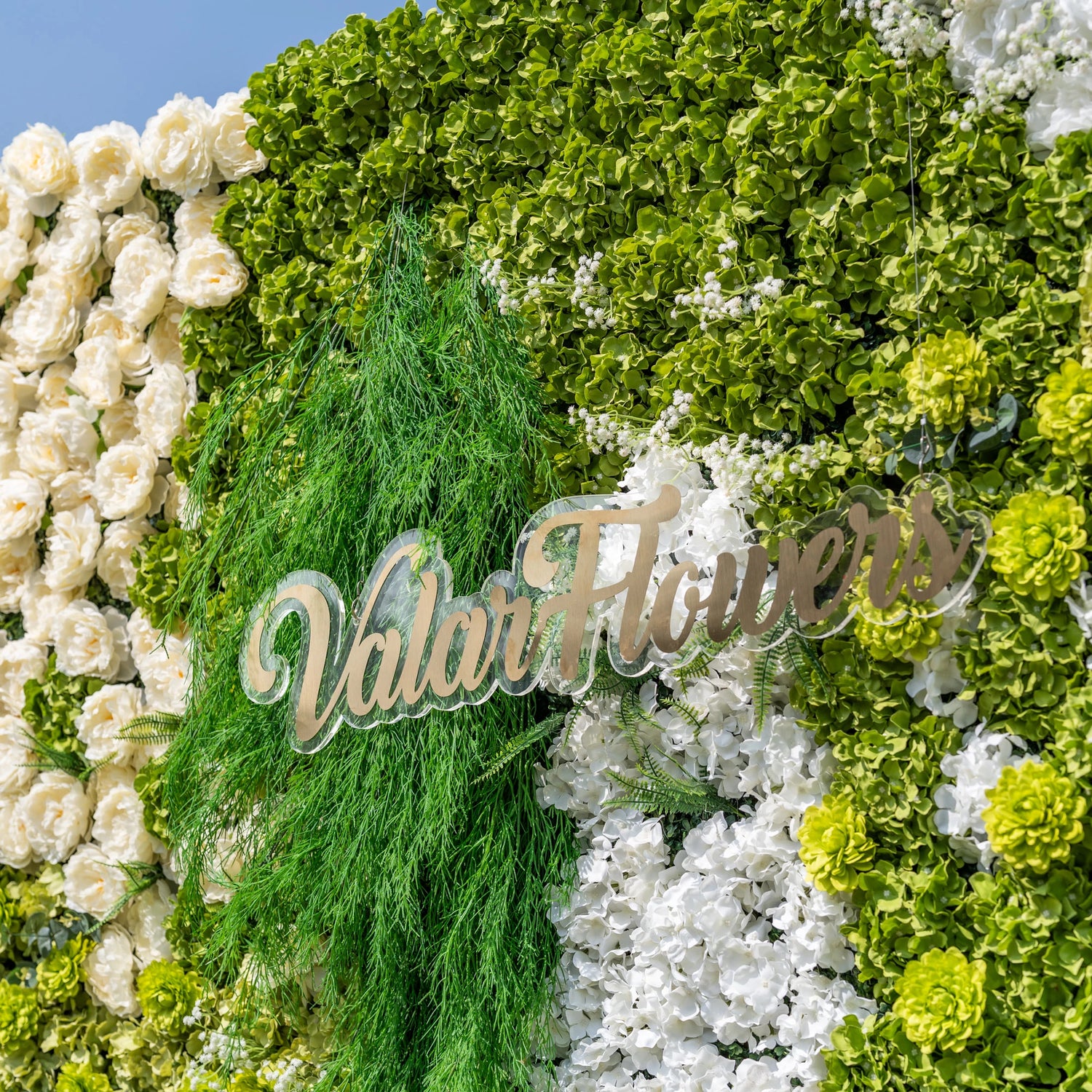 This close-up highlights Valar Flowers’ premium artificial flower wall, featuring lush green hydrangeas, cascading foliage, soft white blooms, and ivory roses, with a sleek metallic “ValarFlowers” logo sign. High-quality, hyper-realistic petals and dense, layered greenery create a lifelike, maintenance-free display, ideal for wedding backdrops, photo booths, and luxury events, offering a fresh, elegant aesthetic for professional decor.