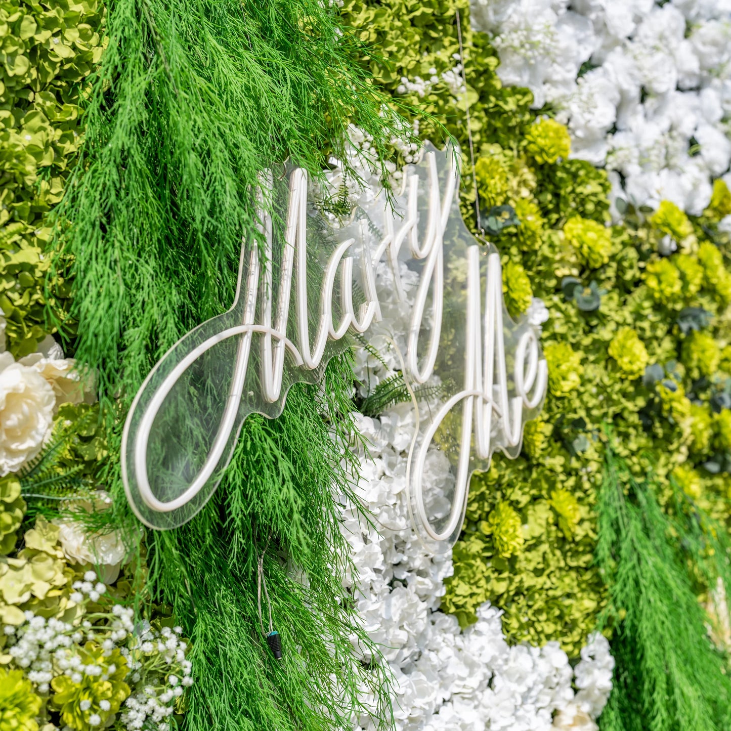 This close-up showcases a glowing white "Marry Me" neon sign mounted on Valar Flowers’ premium artificial floral wall backdrop. The sign is framed by cascading green foliage, lush green hydrangeas, and soft white blooms, creating a romantic focal point. Ideal for proposals, weddings, and luxury events, this combination of neon lighting and hyper-realistic, maintenance-free flowers offers an elegant, nature-inspired aesthetic perfect for photo booths and special occasion decor.