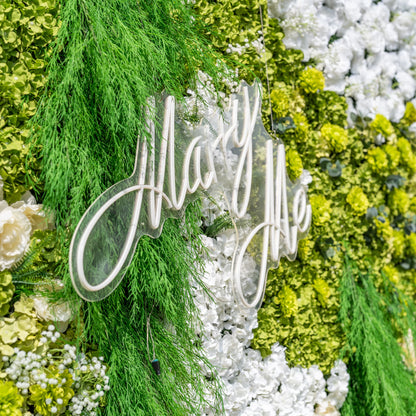 This close-up showcases a glowing white "Marry Me" neon sign mounted on Valar Flowers’ premium artificial floral wall backdrop. The sign is framed by cascading green foliage, lush green hydrangeas, and soft white blooms, creating a romantic focal point. Ideal for proposals, weddings, and luxury events, this combination of neon lighting and hyper-realistic, maintenance-free flowers offers an elegant, nature-inspired aesthetic perfect for photo booths and special occasion decor.