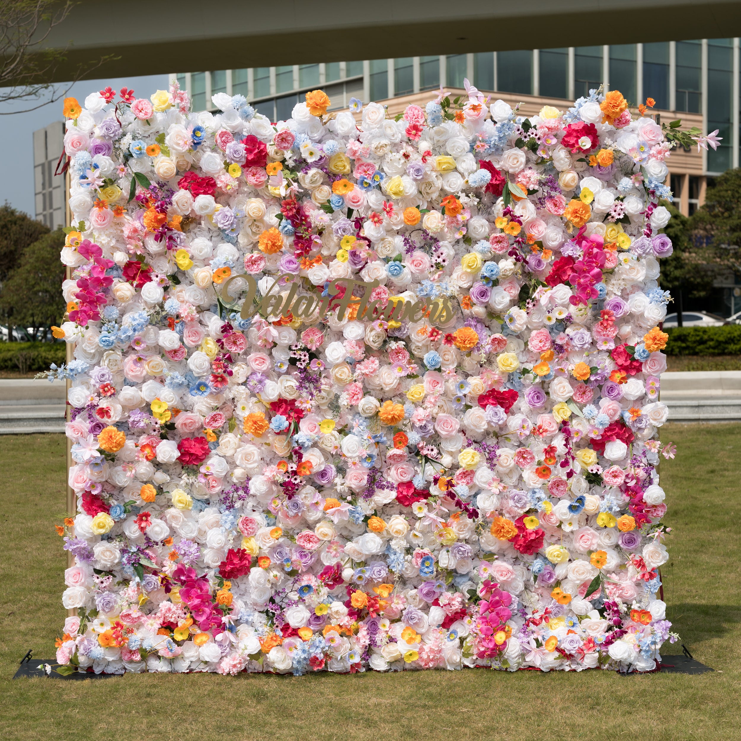 Valar Flowers premium vibrant mixed artificial flower wall backdrop, a dense, lush floral panel bursting with a rainbow of lifelike blooms—roses, poppies, carnations, orchids, and filler flowers in pink, orange, yellow, blue, purple, and white. This stunning, romantic backdrop is perfect for outdoor weddings, proposals, engagement parties, photo booths, and luxury event decor.