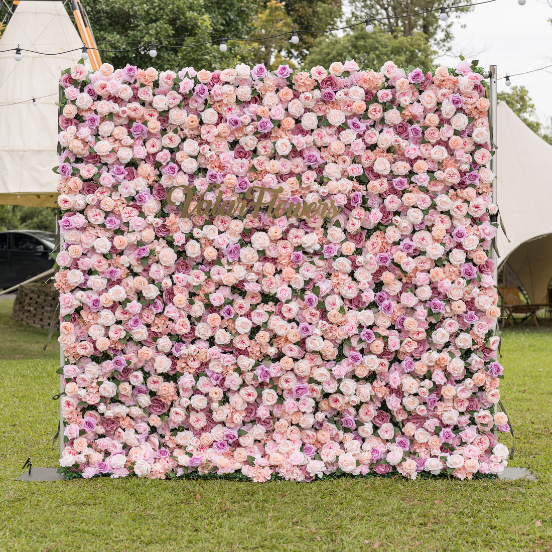 Three-quarter angle of Valar Flowers Sunset Romance artificial flower wall showcasing layered pink peach and lavender roses with ivory hydrangeas outdoors against white tents and string lights for enchanting garden wedding ceremonies and bridal showers
