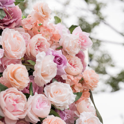 Side-angled close-up of Valar Flowers Sunset Romance wall with metallic script sign among pink and peach roses with shallow focus blurring into greenery and sky for sophisticated salon boutique and bridal suite interior decor installations