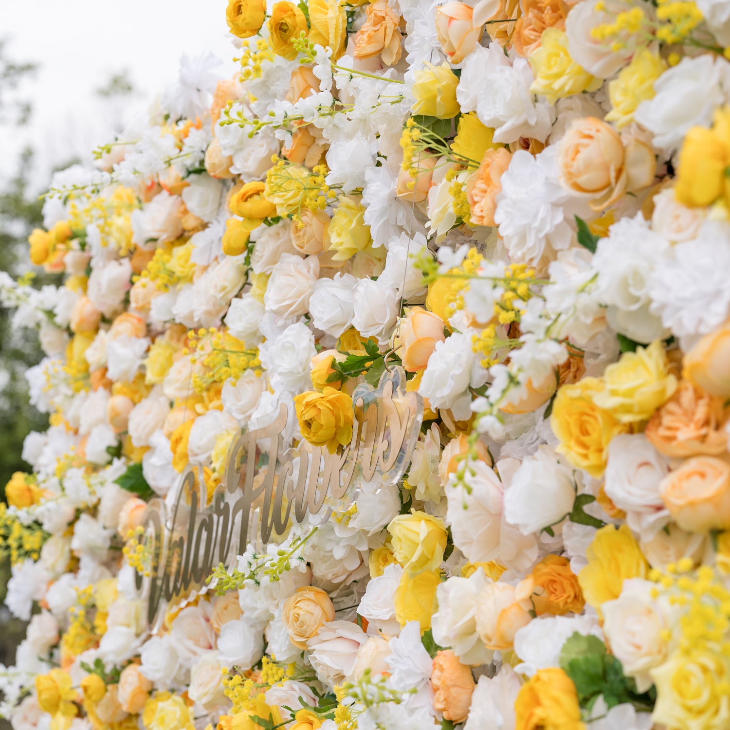 Side-angled close-up of Valar Flowers wall: metallic script sign among buttery yellow and cream roses with blurred greenery background. Cheerful sophistication for bridal showers, birthdays, and garden event decor.