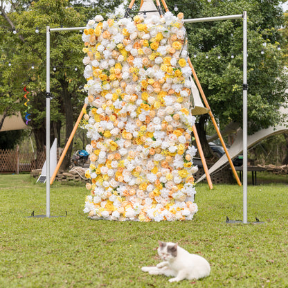 Valar Flowers Sun-Kissed Meadow wall on green lawn: dense white and yellow roses on silver frame with lounging cat in foreground and tents beyond. Whimsical charm for outdoor garden weddings and celebration photography.